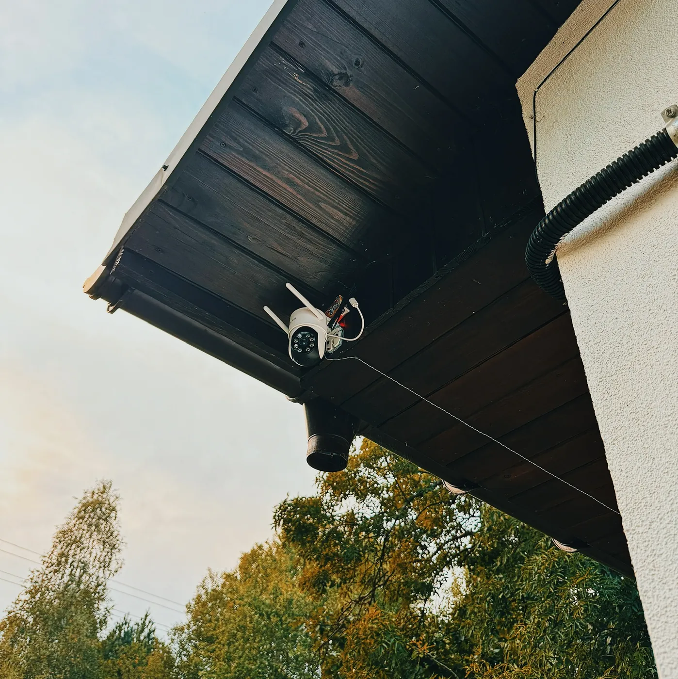 Seen from below: a small white pan-tilt camera with two antennas under a dark wooden eave, a black corrugated conduit running down the white rendered wall beside it, trees still in full leaf and pale sky behind