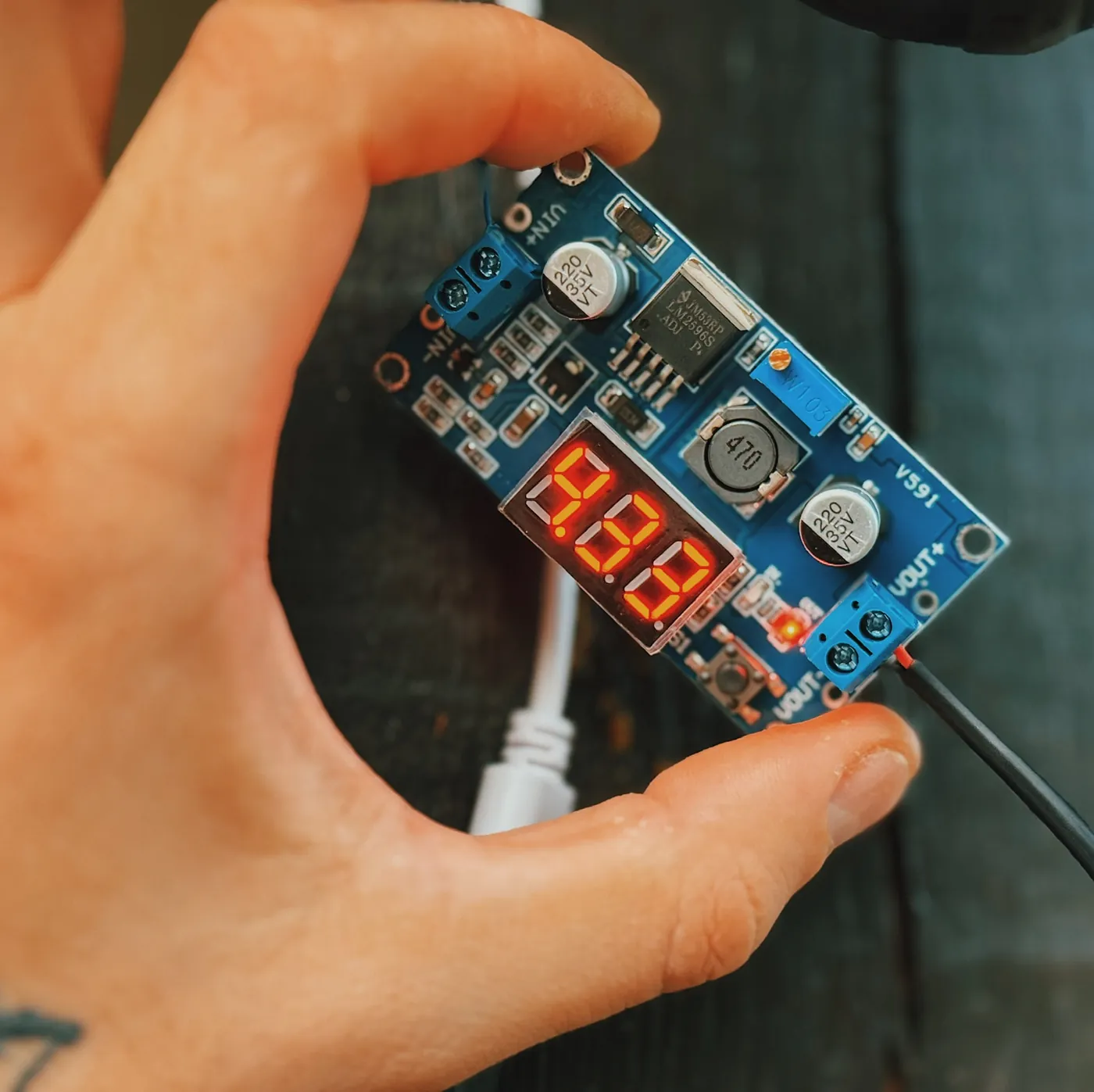 A hand holding a small blue step-down board marked LM2596S-ADJ, with two capacitors, an inductor, a blue adjusting screw and a lit red three-digit display, a black and a red lead screwed into the output terminal