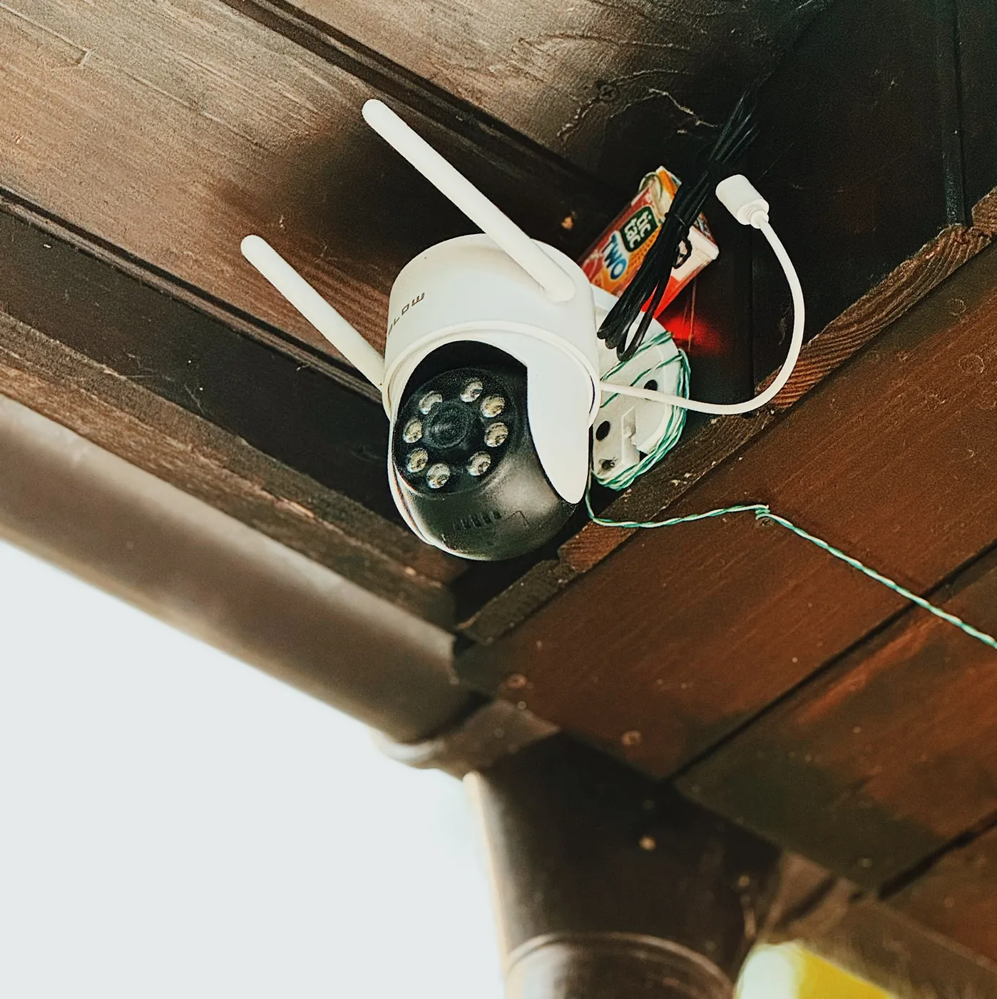 A white outdoor pan and tilt camera with two aerials mounted under a brown roof eave, the same Tic Tac box zip-tied to the timber beside it, a white power lead and a green twisted conductor running away out of shot
