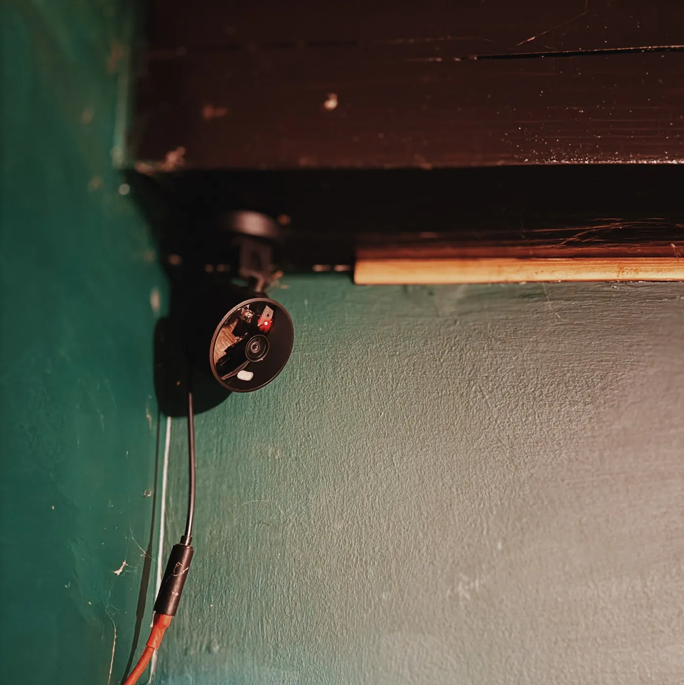 A round black indoor camera on a small bracket in the corner under a dark wooden beam, on a deep green wall, one cable running down from it to an inline connector where a grey lead becomes an orange one