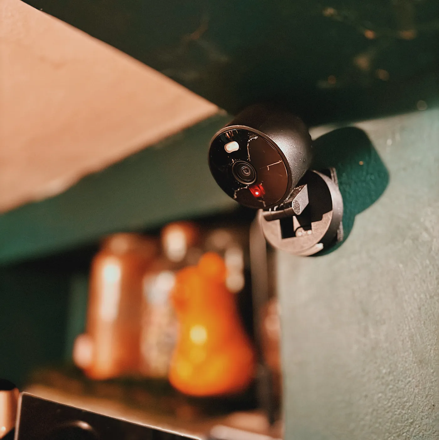 A black cylindrical indoor camera on a wall bracket beside a pale shelf, a red light lit under its lens, out-of-focus jars and warm light on the shelf behind it